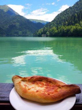 Güzel Abhazya Gölü Ritsa ve dağlarının arka planında beyaz bir tabakta sıcak Adjarian Khachapuri. Ulusal Kafkas yemekleri. Lezzetli aperatif ve muhteşem manzara.