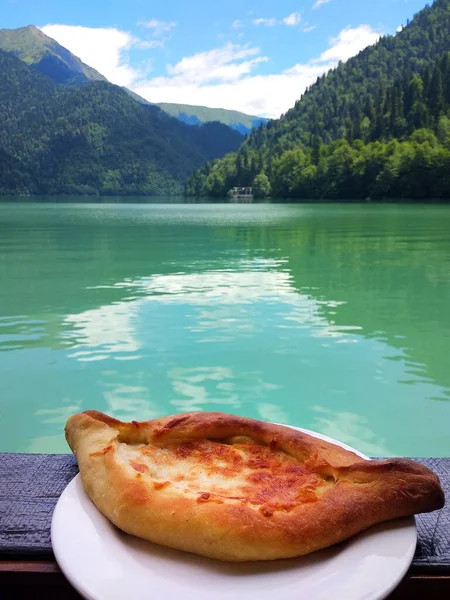 Güzel Abhazya Gölü Ritsa ve dağlarının arka planında beyaz bir tabakta sıcak Adjarian Khachapuri. Ulusal Kafkas yemekleri. Lezzetli aperatif ve muhteşem manzara.
