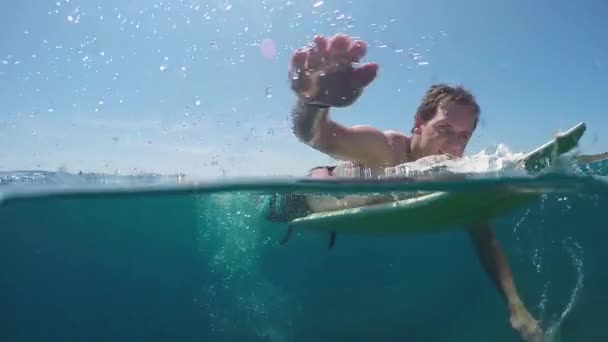 SLOW MOTION UNDERWATER: Smiling surfer man paddling on a surf in vast blue sea — Stock Video