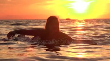 Ağır çekim: okyanusta altın günbatımında sörf tahtası üzerinde kürek çekmeye güzel kız