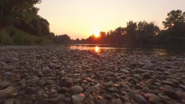 AERIAL, CLOSE UP: Stony riverbank and water surface reflecting golden sky — Stock Video