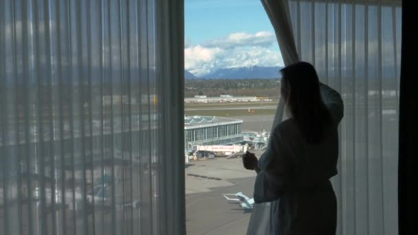 Молодая туристка в халате двигает занавеску и наблюдает за аэропортом. — стоковое видео