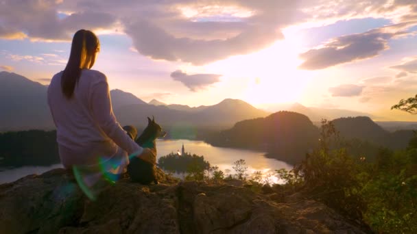 FECHAR UP: Menina sentada na rocha e assistindo o pôr do sol acima Bled com cão. — Vídeo de Stock