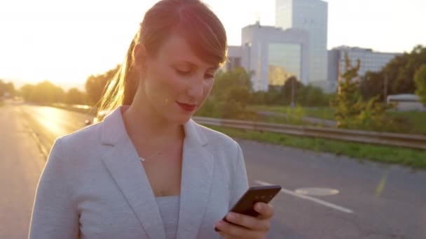 閉じる:若いビジネスマンの女性が歩道を歩いて日没時にテキストメッセージを送る. — ストック動画