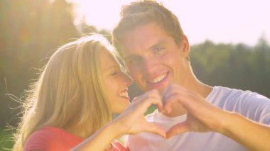 CLOSE UP: Blonde woman kisses man on the cheek while holding up a heart shape.