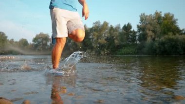 LOW ANGLE: Glassy water droplets fly towards camera as jogger runs in the stream