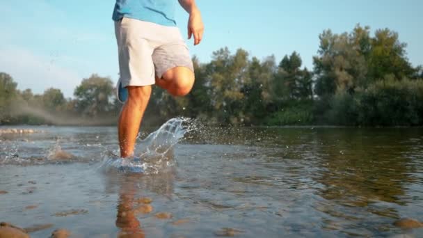 Niedriger Winkel: Glasartige Wassertröpfchen fliegen auf Kamera zu, als Jogger im Bach rennt — Stockvideo