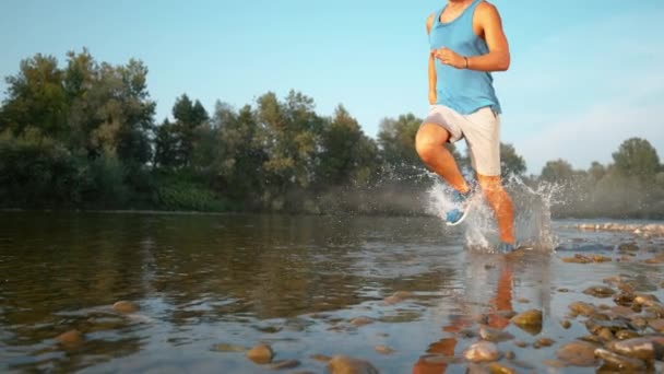 ÁNGULO BAJO: Joven feliz corriendo en el río y salpicando agua refrescante . — Vídeos de Stock