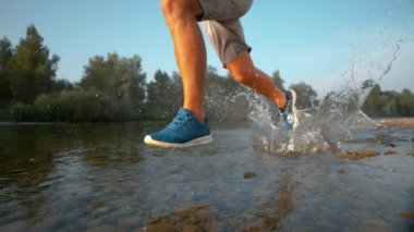 LOW ANGLE: Cinematic shot of young male runner's legs splashing the stream water