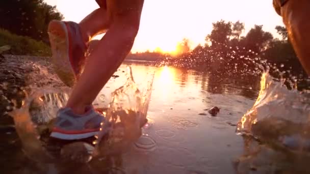 FLARE DE LA LENTE: Pareja irreconocible corriendo en un arroyo poco profundo y hacia el atardecer . — Vídeos de Stock