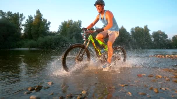 SUPER SLOW MOTION: Athletic cross country biker riding along a shallow stream. — ストック動画