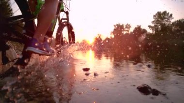 SLOW MOTION: Sportsman riding his bicycle in the shallow stream towards sunset.