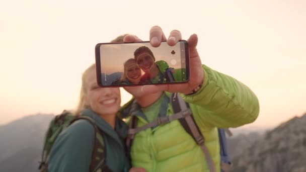 CERRAR: Sonriente pareja de jóvenes excursionistas tomando una selfie en la cima de la montaña al amanecer . — Vídeo de stock