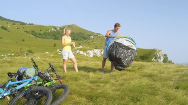 MOVIMIENTO Lento Pareja alegre y activa instalando una tienda de campaña en el prado en una montaña — Vídeo de stock