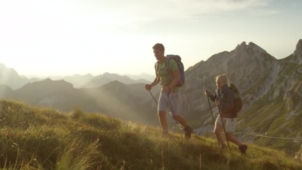 LENS FLARE: Goldene Sonnenstrahlen scheinen auf junges Paar, das einen steilen Hügel in den Alpen hinaufwandert — Stockvideo