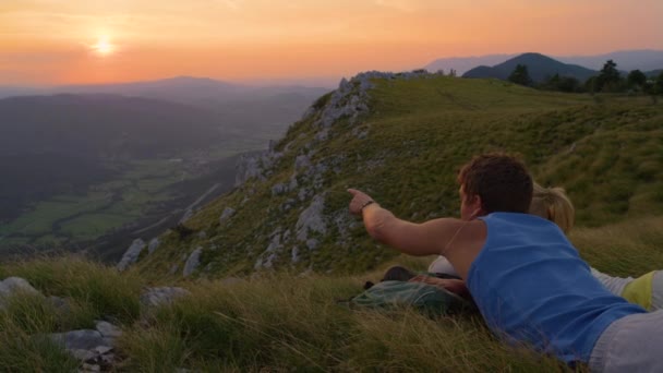 Мужчина, указывающий на горы своей девушке, пока они лежат в траве . — стоковое видео