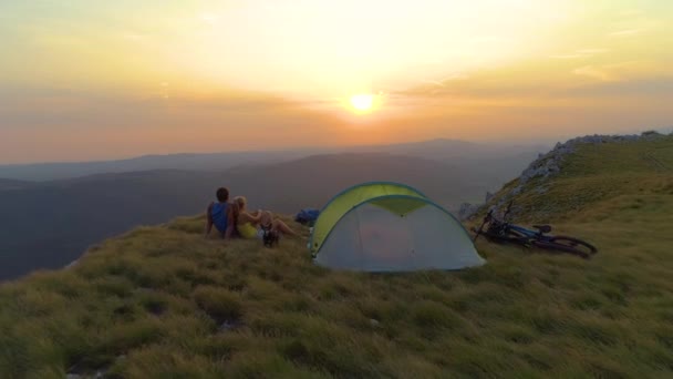 AÉRIEN : Les rayons du soleil doré brillent sur le chiot et le couple câlinant par tente au coucher du soleil . — Video