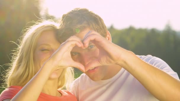 Мужчина целует подружку в щеку, когда они делают знак сердца пальцами . — стоковое видео
