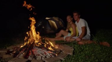 SLOW MOTION: Cheerful man strikes campfire with stick while snuggling with girl.
