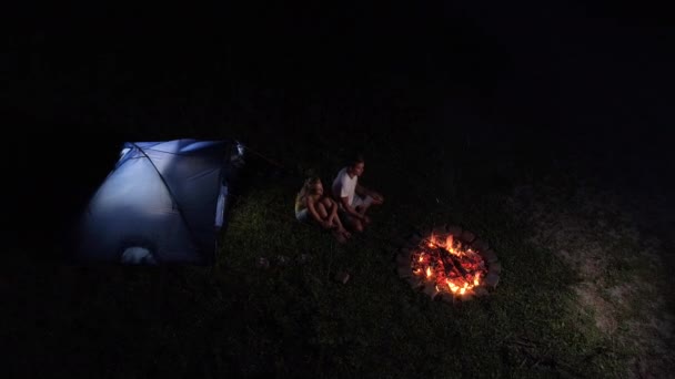 AERIAL: Volando por encima de una joven pareja sentada junto al fuego durante un viaje de campamento . — Vídeo de stock