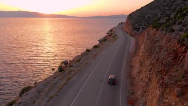 DRONE: Carro desce estrada vazia que leva ao longo do penhasco e mar calmo ao pôr do sol — Vídeo de Stock