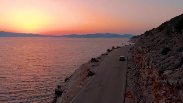 Donker gekleurde auto rijdt langs het rotsachtige terrein en kalme zee bij zonsondergang. — Stockvideo