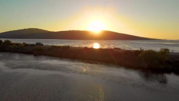 AERIAL: Lecąc w kierunku zachodzącego słońca oświetlającego morze i samochód płynący ulicą — Wideo stockowe