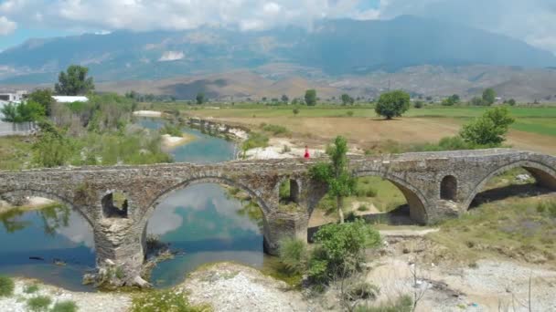 DRONE Ung kvinna i röd klänning promenader längs den antika ottomanska bron i Albanien — Stockvideo