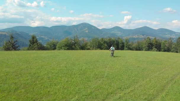 AERIAL: Flying along a grassy trail leading a mountain biker across the meadow. — Stock Video