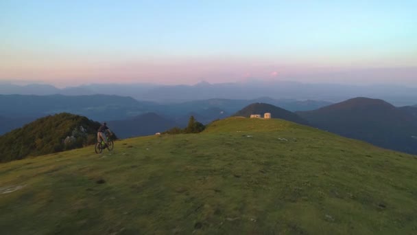 AERIAL: Extreme mountainbiker snelheden langs een smal graspad bij zonsondergang. — Stockvideo