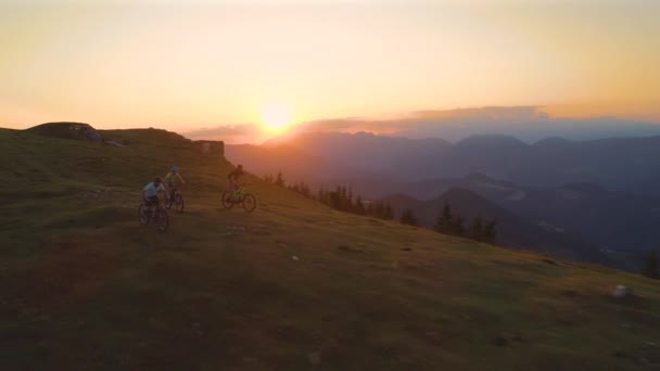 AERIALE: Tre ciclisti in mountain bike che scendono velocemente da una collina erbosa al tramonto dorato . — Video Stock