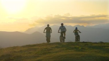 Güneşli bir akşamda üç sportif turist çayırda elektrikli bisiklet sürüyor..