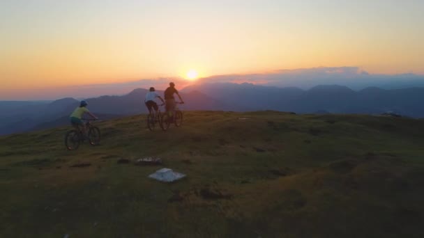 Volando sobre tres ciclistas de montaña bajando por una colina cubierta de hierba al atardecer . — Vídeos de Stock