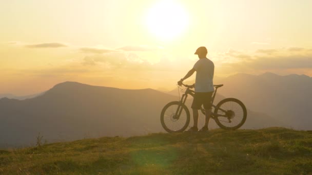 MOCIÓN LENTA: Joven turista masculino observa el paisaje antes del ciclismo de montaña — Vídeos de Stock