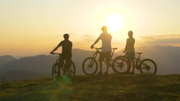SILHOUETTE: Los ciclistas de montaña descansan en la cima de la montaña cubierta de hierba y observan el amanecer . — Vídeos de Stock