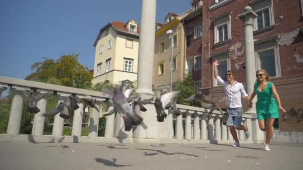 Nízký úhel hravý turistický pár strašit holuby při chůzi přes most — Stock video