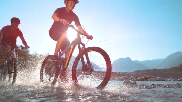 LÅNGA: Vattendroppar flyger runt två killar rider elektriska cyklar längs floden. — Stockvideo