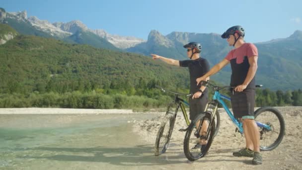 SLOW MOTION: Mladí cyklisté odpočívají u řeky a pozorují krásné hory — Stock video