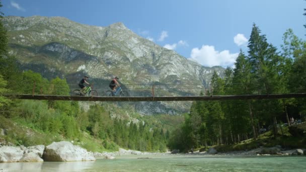 LÄGG: Två mountainbikers rider över en hängbro ovanför lugna floden. — Stockvideo