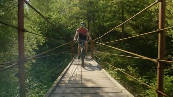FLARE DE LA LENTE: Hombre alegre monta una bicicleta a través de un puente colgante en un día soleado. — Vídeo de stock