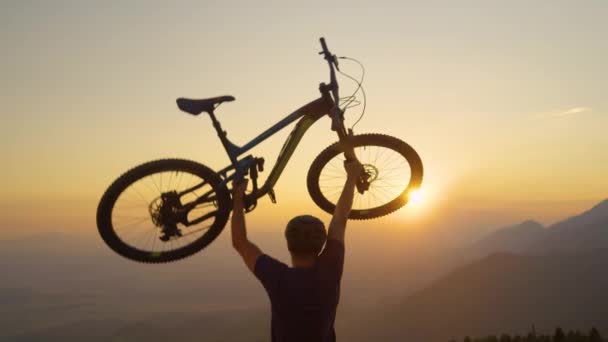 LOSE UP: Oigenkännlig man lyfter sin cykel i luften vid gyllene solnedgång. — Stockvideo