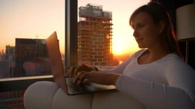 Genç bir kadın, altın güneş ışınları odasına girerken dizüstü bilgisayarı üzerinde çalışıyor.