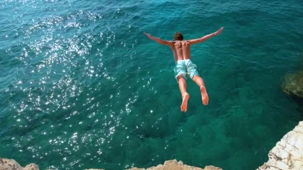 COPY SPACE: Man jumping off a rocky ledge and into the glistening blue ocean. — Stock Video