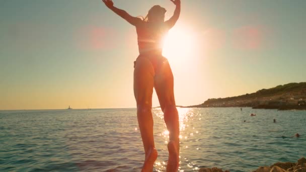 FLARE DE LA LENTE: La chica salta de un acantilado y patea las piernas mientras se sumerge en el océano. — Vídeos de Stock