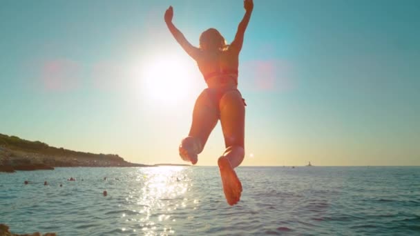 SUN FLARE: dziewczyna skacze z klifu i kopie nogi z powrotem podczas nurkowania w oceanie. — Wideo stockowe