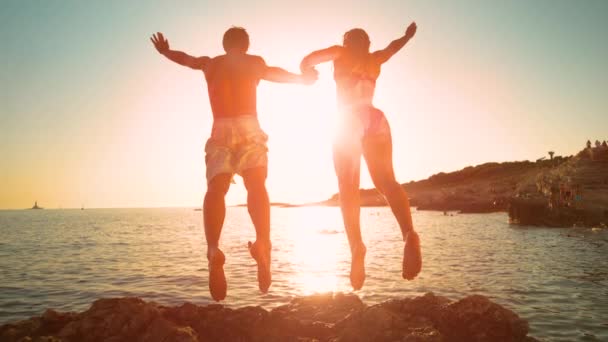 LENS FLARE: Cheerful couple on summer vacation jumps into the ocean at sunrise. — Stock Video