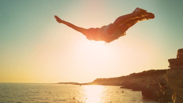 CERRAR: La puesta de sol dorada brilla en el buzo de roca saltando de un acantilado y en el mar. — Vídeos de Stock