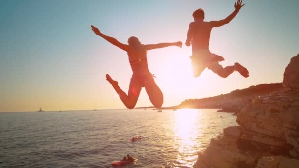 FLARE DE LA LENTE: La mujer sostiene la mano de los novios mientras salta de un acantilado hacia el mar. — Vídeos de Stock