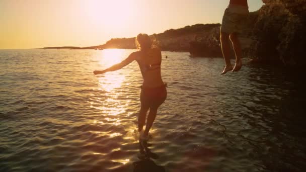 FLARE DE LA LENTE: Pareja joven salta al mar Mediterráneo en una soleada noche de verano — Vídeos de Stock