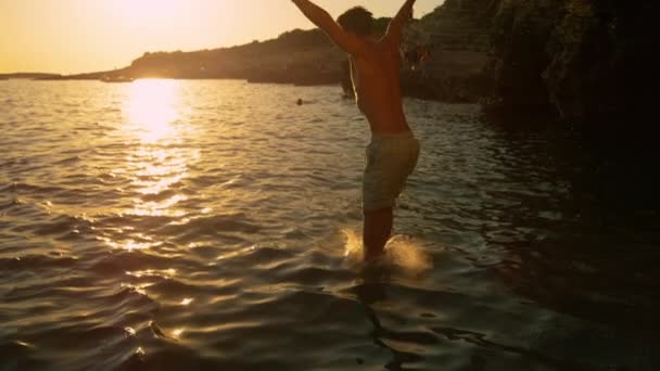SUN FLARE Glad man med utsträckta armar hoppar fötterna först i havet vid solnedgången — Stockvideo